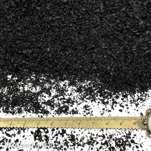 Op de afbeelding: Een verzameling kleine, onregelmatige zwarte mineraalkorrels verspreid over een wit oppervlak, geplaatst boven een messingkleurige metalen liniaal die tot 10 centimeter meet, met de tekst Makers Minerals zichtbaar onderaan.