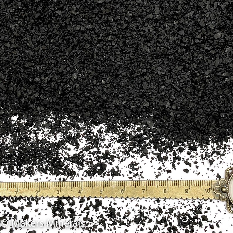 Op de afbeelding: Een verzameling kleine, onregelmatige zwarte mineraalkorrels verspreid over een wit oppervlak, geplaatst boven een messingkleurige metalen liniaal die tot 10 centimeter meet, met de tekst Makers Minerals zichtbaar onderaan.