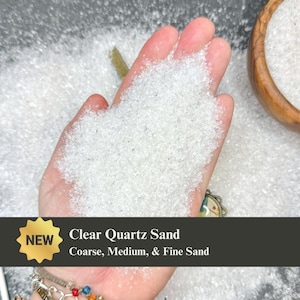 May include: A pile of clear quartz sand with a crystalline texture held in a palm. The text on the image reads NEW Clear Quartz Sand Coarse, Medium, & Fine Sand. The sand consists of small, translucent, jagged particles that reflect light.