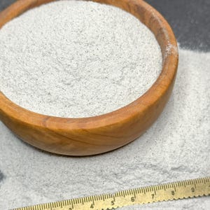 May include: A wooden bowl containing a light grey, fine powder. The bowl has a natural wood grain and sits on a surface with scattered powder. A ruler is placed near the bowl, providing a size reference.