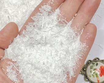 May include: A collection of small, translucent, needle-like white crystals piled in a palm. The crystals have a sharp, elongated, and splintered appearance. In the lower corner, a round bronze-toned pendant features a floral design with red petals.