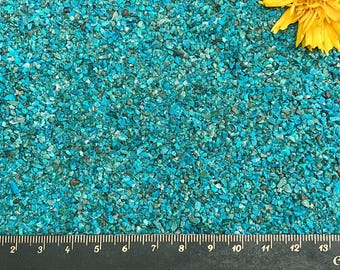 Può includere: Una collezione di piccole pietre frantumate color turchese sparse su una superficie, accompagnate da un fiore giallo brillante nell'angolo in alto a destra e da un righello nero che misura da 2 a 13 cm lungo il bordo inferiore.