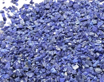 May include: A collection of small, crushed lapis lazuli stones featuring a deep, vibrant blue color with scattered flecks of white and grey mineral inclusions. The fragments are irregular in shape and size, creating a textured, granular appearance.