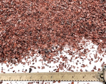 May include: A collection of small, irregular, dark red crushed stone fragments scattered across a surface. A brass-colored metal ruler is positioned at the bottom, showing a scale in centimeters with markings from 1 to 10 clearly visible.