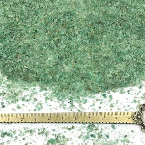 May include: A collection of small, irregular green mineral fragments scattered across a white surface. A brass-colored metal ruler is positioned horizontally at the bottom, showing measurements in centimeters from 1 to 10, with a decorative floral end.