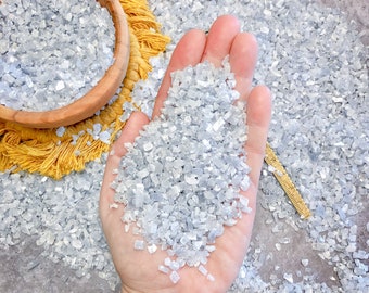Può includere: Una mano che tiene un mucchio di piccoli cristalli blu iridescenti trasparenti. I cristalli sono sparsi su una superficie grigia.