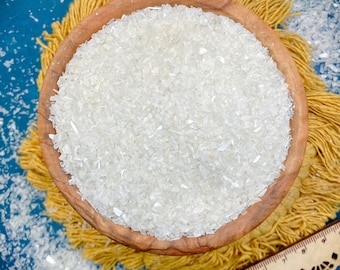 May include: A round wooden bowl filled with small, translucent white crushed glass fragments. A wooden ruler is visible at the bottom right, showing a measurement scale in inches. The bowl rests on a yellow fringed mat against a blue background.