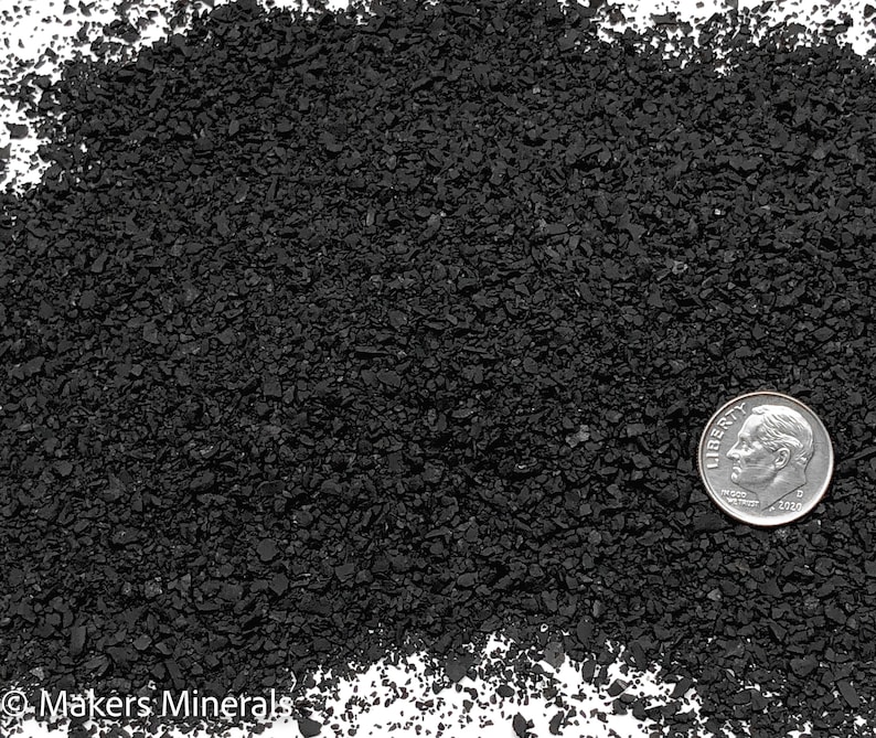 Op de afbeelding: Een hoop kleine, grove, zwarte mineraalkorrels met een onregelmatige, grillige textuur. Een zilverkleurige Amerikaanse munt van tien cent, met een diameter van ongeveer 1,8 cm, is aan de rechterkant geplaatst voor schaal. De tekst Makers Minerals is zichtbaar in de linkerbenedenhoek.