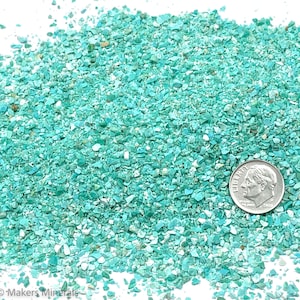 Può includere: Primo piano di un mucchio di trucioli di turchese. I trucioli sono di varie tonalità di blu e verde, e alcuni sono iridescenti. Una moneta da dieci centesimi di euro d'argento è posizionata sopra i trucioli per dare un'idea della scala.