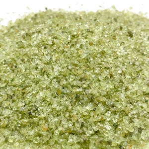 May include: Close-up of a pile of small, light green peridot gemstone chips. The chips are irregularly shaped and have a translucent quality, with varying shades of green. The gemstones are scattered, creating a textured surface.