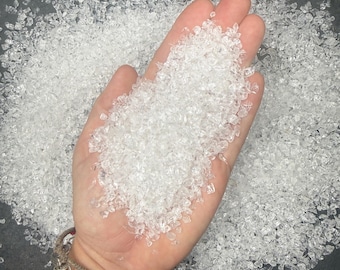 May include: A large handful of small, clear, crushed glass fragments with irregular shapes and sharp edges. The translucent pieces are piled high in the palm, reflecting light and appearing like tiny, jagged diamonds or clear quartz crystals.