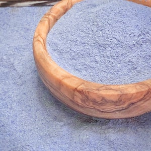Op de afbeelding: Een houten kom gevuld met fijn, lichtpaars poeder. De kom is gemaakt van lichtbruin hout met zichtbare nerfpatronen. Het poeder morst op het oppervlak en creëert een textuur, korrelig uiterlijk. De afbeelding is goed verlicht en benadrukt de kleur en textuur.