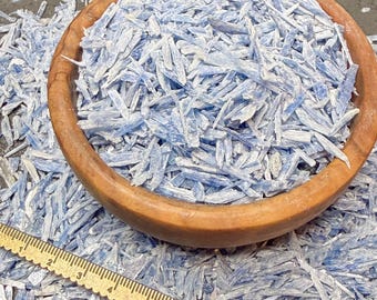 Puede incluir: Un cuenco de madera redondo lleno de pequeños fragmentos alargados de cristales de cianita azul claro. Una regla de metal está colocada en primer plano, mostrando que los cristales miden aproximadamente 18 cm de largo en la escala visible.