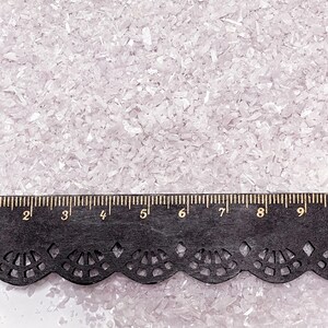 May include: A collection of small, translucent, pale pink crushed glass fragments spread evenly. A black ruler with a decorative scalloped edge is positioned across the bottom, showing measurements in inches from two to nine inches.