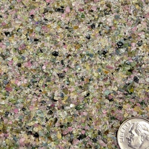 May include: A collection of small, crushed mineral fragments in shades of pink, green, yellow, blue, and black. A United States ten-cent coin is positioned in the bottom right corner to provide a sense of scale for the tiny, irregular stone pieces.