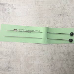 May include: Two knitting needles, size 19, are shown on a light green card. The needles are thin, silver-coloured, and have a dark grey ball at one end. The card has text that reads "Knitting Needles Size 19 (1.0)" and "Sharonsminiknits".