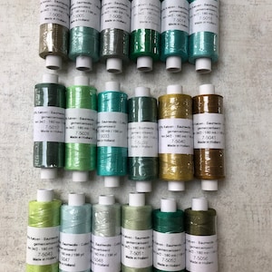 Op de afbeelding: Een assortiment katoenen garenspoelen in verschillende groentinten, van licht mintgroen tot diep bosgroen. Elke spoel heeft een wit etiket met zwarte tekst en is ongeveer 180 meter lang.