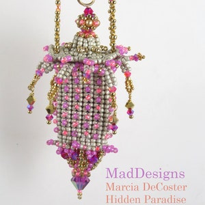 Könnte beinhalten: Ein Perlenanhänger mit einem rosa-weißen Design, goldenen Akzenten und einem Kristalltropfen. Der Anhänger ist mit "MadDesigns Marcia DeCoster Hidden Paradise" beschriftet.