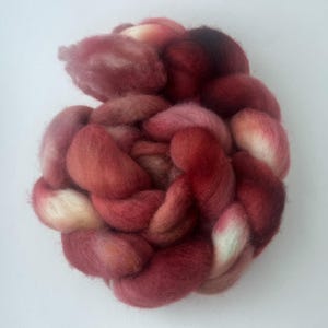 May include: A braided bundle of soft, fluffy wool roving featuring a variegated color palette that transitions through shades of deep burgundy, dark crimson, dusty rose, and creamy off-white, arranged in a thick, textured, circular coil.