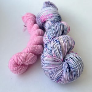 Könnte beinhalten: Zwei Knäuel Garn werden vor einem weißen Hintergrund gezeigt. Eines ist einfarbiges, helles Rosa, das andere ein mehrfarbiges Garn mit Rosa-, Blau- und Weißtönen. Das mehrfarbige Garn hat kleine Farbflecken.