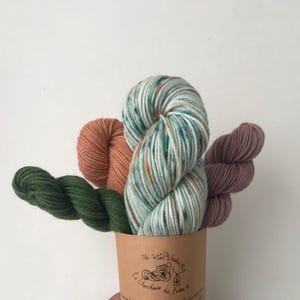 May include: Three skeins of yarn in a paper cup. The yarn is green, pink, and a light blue with orange flecks. The cup has a label that reads "The Wool Studio, La Chevrehouse de l'Ours".