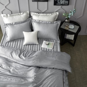 May include: A bed with a gray comforter and pillows. The bed has a gray quilted bedspread, two white pillows, and four gray pillows. A small black side table holds a vase of flowers, a mug, and a clock.