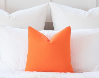 Op de afbeelding: Een feloranje vierkant kussen ligt op een bed en contrasteert met het witte beddengoed en de kussens. Het kussen is het middelpunt, met een effen, levendige kleur. Het bed is bedekt met een witte, zachte deken.