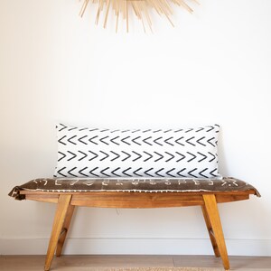 May include: A wooden bench with a brown patterned cushion and a white lumbar pillow with a black chevron pattern. The bench is in front of a white wall with a woven sunburst wall hanging.