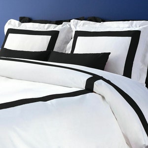 May include: White bedding with black trim. The bedding has a simple, modern design with a black border around the edges of the duvet cover and pillowcases.