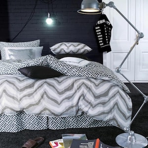 Peut inclure: Une couette à motif chevron gris et blanc sur un couvre-lit à motif noir et blanc. Le lit est décoré d'oreillers décoratifs à motif noir et blanc et d'un oreiller décoratif argenté et noir. Un calendrier noir et blanc est accroché à une porte blanche derrière le lit. Une lampe argentée avec un cordon noir est posée sur le sol à côté du lit. Trois livres sont posés sur le sol devant le lit. Les livres ont les mots "HOME" et "NOTHING THAT YOU DO NOT LOVE" imprimés sur les couvertures.