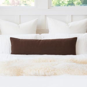 May include: A long, rectangular brown pillow rests on a white bed, with white pillows in the background. A cream-colored, fluffy throw is at the foot of the bed. The scene is bright and airy, suggesting a bedroom setting.