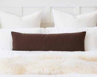 May include: A long, rectangular brown pillow rests on a white bed, with white pillows in the background. A cream-colored, fluffy throw is at the foot of the bed. The scene is bright and airy, suggesting a bedroom setting.