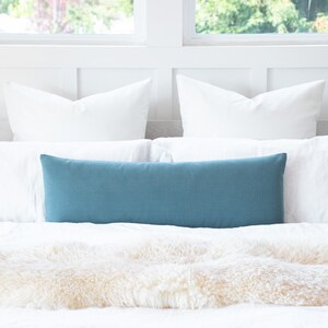 May include: A teal rectangular lumbar pillow rests on a bed with white pillows and a fluffy, cream-colored throw. The pillow is centered on the bed, providing a pop of color against the neutral bedding. The room has a bright, airy feel.