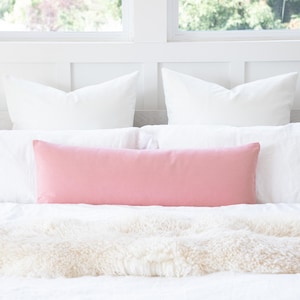 Pode incluir: Uma cama é feita com travesseiros e roupa de cama brancos. Uma almofada longa e retangular rosa claro repousa sobre a roupa de cama branca. Uma manta de pele sintética branca e fofa está aos pés da cama.