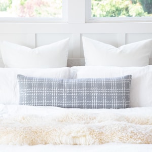 Pode incluir: Uma cama com almofadas brancas e uma almofada lombar cinza com um padrão xadrez branco. A cama é coberta com um lençol branco e uma manta de pele sintética creme. O fundo inclui uma cabeceira branca e janelas.