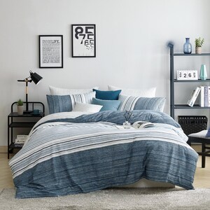 May include: A bed with a blue and white striped duvet cover and matching pillows. The room has a minimalist design with framed art, a black side table, and a gray rug. A black lamp and a shelf with decorative items are also visible.