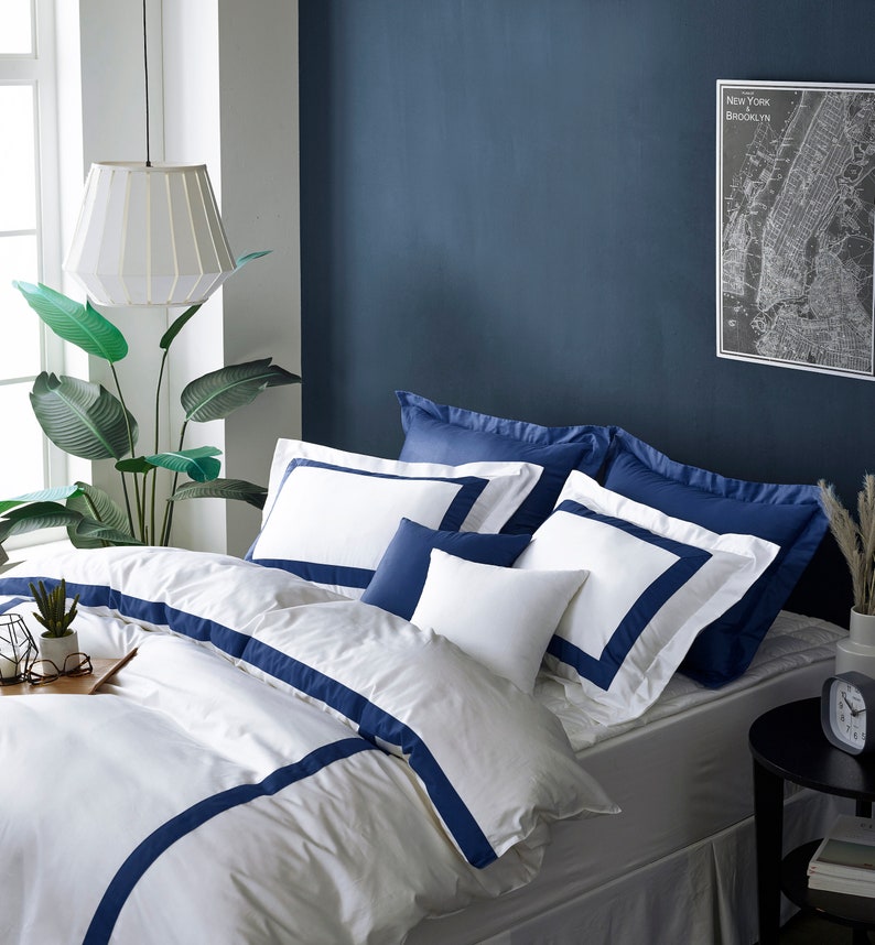 Pu&ograve; includere: Biancheria da letto bianca con bordino blu navy, un copripiumino bianco con strisce blu navy e un set di federe bianche e blu navy. La biancheria da letto &egrave; su un letto con un materasso bianco e un copriletto bianco. C'&egrave; una pianta in un vaso sul lato sinistro del letto e un orologio su un comodino nero sul lato destro del letto.