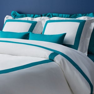 Può includere: Biancheria da letto bianca con bordino blu turchese. La biancheria da letto è disposta su un letto con cuscini blu turchese.