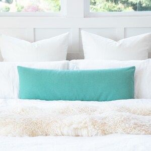 May include: A turquoise blue lumbar pillow on a white bed with a white faux fur throw.
