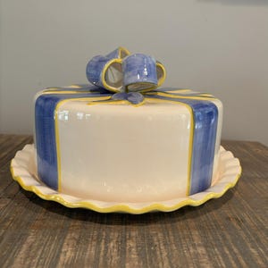 May include: A white ceramic cake plate and cover set, decorated with blue and yellow ribbon and bow details. The plate has a scalloped edge, and the cover is round. The set is designed to resemble a wrapped gift.