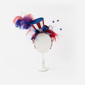 May include: A patriotic headband featuring a miniature top hat with red and white stripes and a blue glitter brim. The headband is adorned with red, white, and blue feathers, a blue bow, and star accents, perfect for Independence Day celebrations.