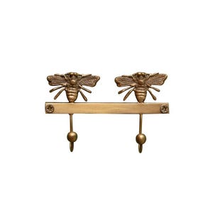 May include: A decorative wall hook featuring two bronze-colored bee figures above two downward-facing hooks. The bees are detailed with wings and body segments, and the hooks have a rounded end.