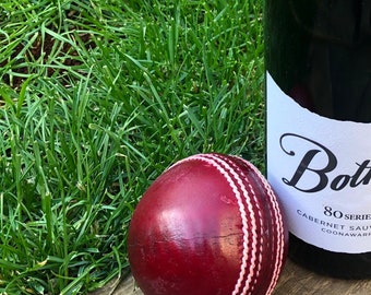 Könnte beinhalten: Ein roter Cricketball mit weißen Nähten liegt auf einer Holzoberfläche neben einer Weinflasche. Das Weinetikett lautet "Bollinger 80 Series Cabernet Sauvignon Coonawarra".