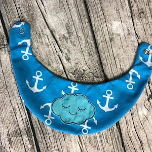 May include: A blue baby bib with a white anchor pattern and a blue cloud with a smiling face. The bib has a snap closure.