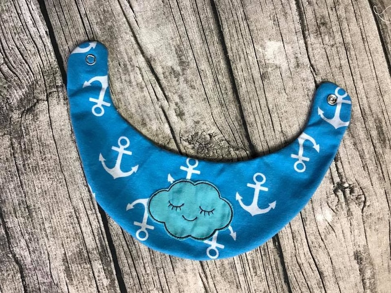May include: A blue baby bib with a white anchor pattern and a blue cloud with a smiling face. The bib has a snap closure.