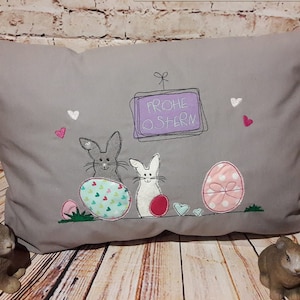 May include: A gray pillow with embroidered bunnies, eggs, and the text "Frohe Ostern" (Happy Easter).