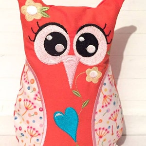 May include: A bright orange owl plush toy with embroidered details. The owl has large black eyes, pink eyelashes, and a blue heart on its chest. The owl is surrounded by a white and pink floral pattern.