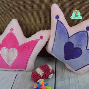 Puede incluir: Dos almohadas de felpa en forma de corona, una rosa con un corazón blanco y la otra morada con un corazón morado.