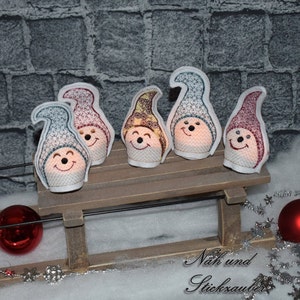 Puede incluir: Cinco pequeños adornos de gnomo de fieltro blanco con caras bordadas y sombreros de varios colores, incluyendo rojo, azul, amarillo y verde. Los gnomos están sentados en un trineo de madera, con un adorno rojo y copos de nieve plateados en el fondo.