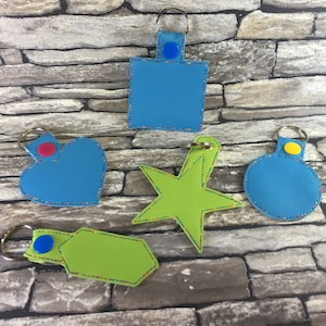 May include: Five different keychains with colorful fabric and stitching. The keychains are shaped like a heart, a star, a circle, and two rectangles. The keychains are all blue, green, and yellow.